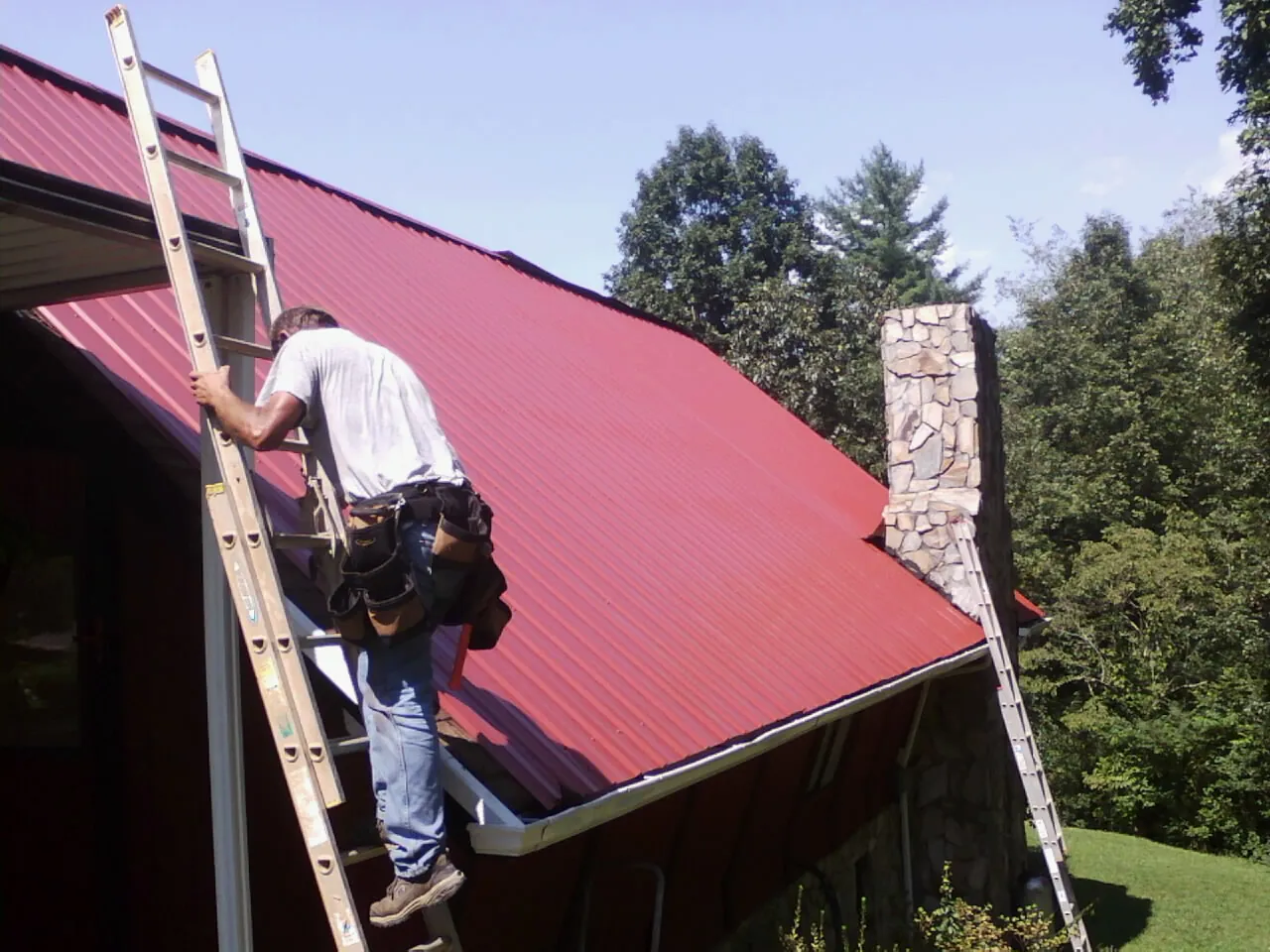 Roofer on a ladder beside a red metal roof and stone chimney — Soffit & Fascia Repair by Roofing Brothers in West Middletown